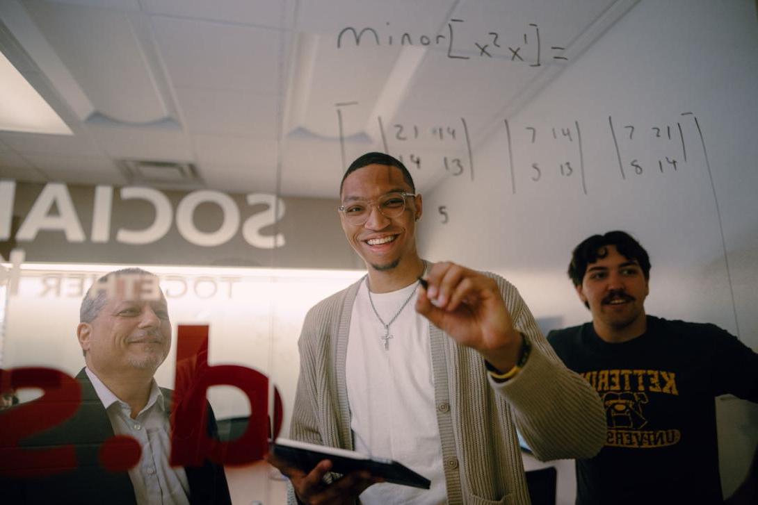 A School of Management Kettering student writes matrix equations on a glass wall. Another student and a professor watch